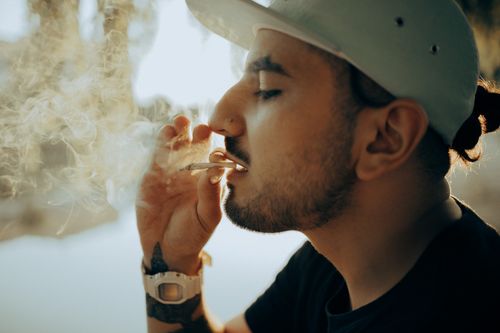 Free Close-up of a man with facial hair and cap smoking outdoors in a casual setting. Stock Photo
