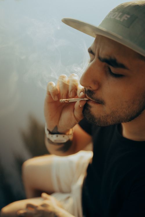 Free A young man with a beard smokes outdoors, wearing a cap and casual clothing. Stock Photo