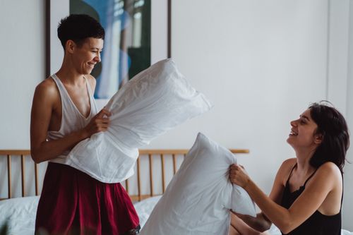Free Joyful same-sex couple enjoying a playful pillow fight together indoors. Stock Photo