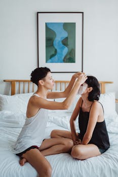 Two women sit on a bed applying facial masks, embracing skincare and relaxation together.