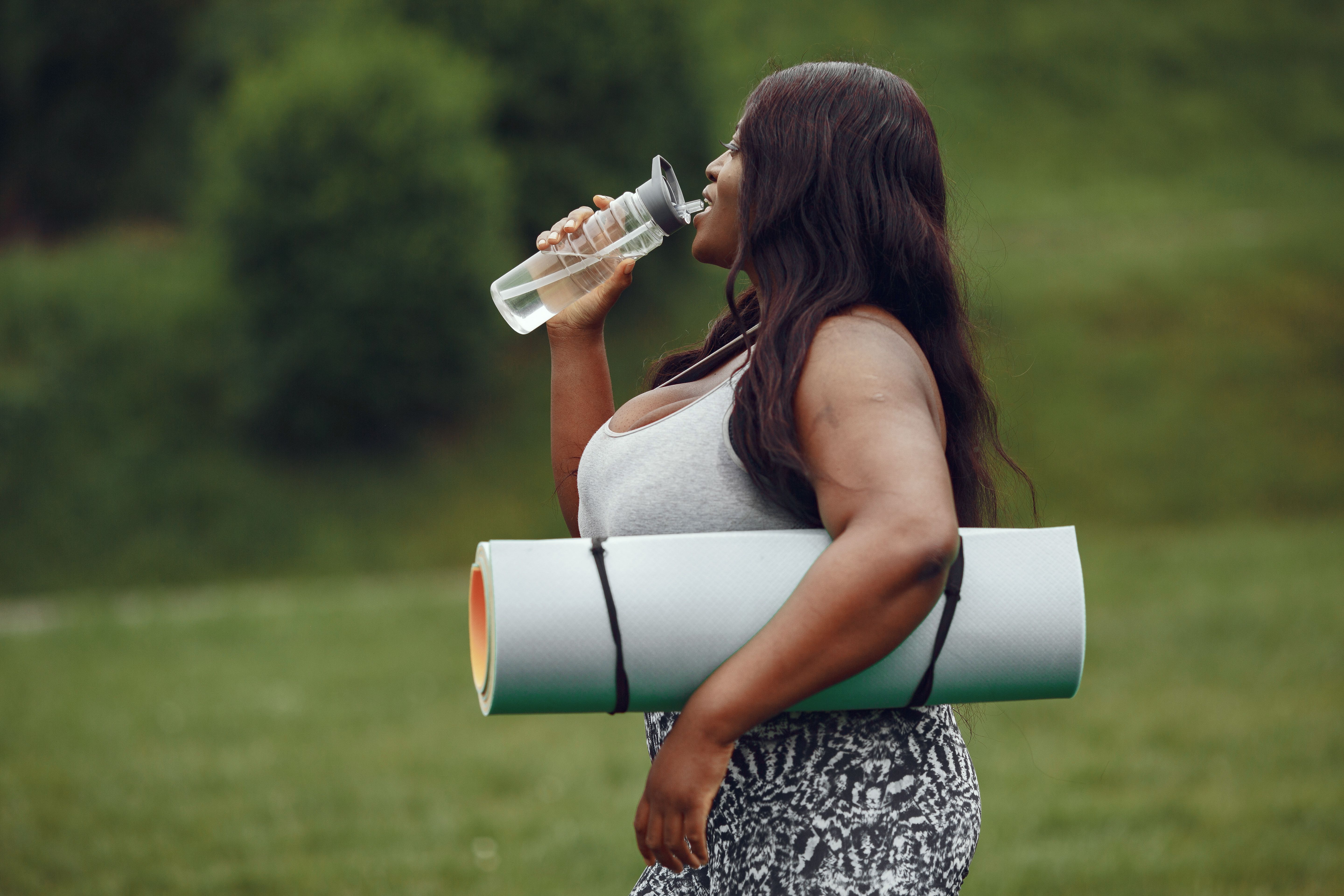 Woman drinking water and carrying yoga mat in green outdoor setting, embracing a healthy lifestyle.