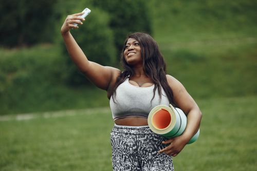 Free Confident black woman takes a selfie while holding exercise mat in a park. Stock Photo