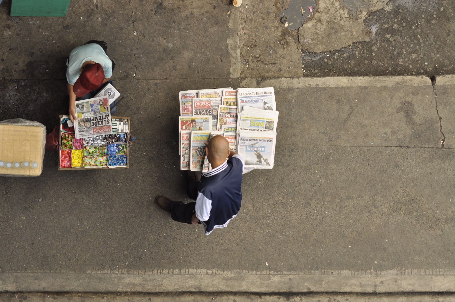 Street vendor arranging newspapers on a concrete sidewalk with candies and public interest articles.