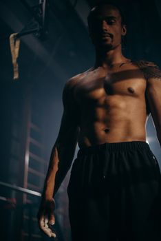 Muscular male athlete standing shirtless in a gym with strong lighting contrasts.