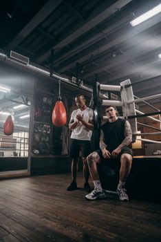 Two muscular men take a break near a punching bag in an industrial-style gym.