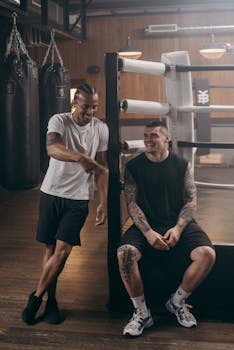 Two muscular athletes enjoying a moment of laughter beside a boxing ring in an indoor gym setting.
