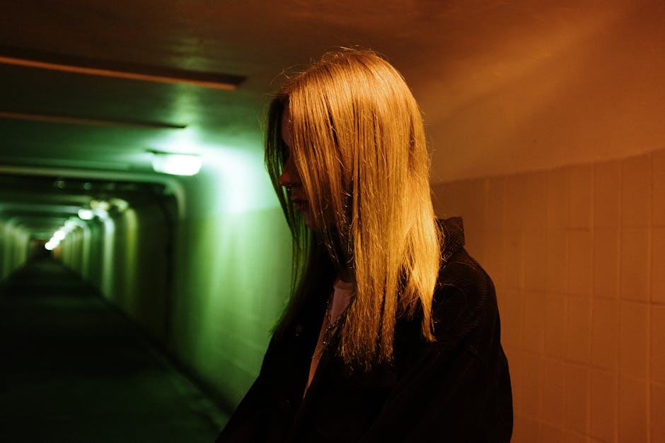 A woman stands in a dimly lit urban underpass with contrasting neon lights casting dramatic shadows.