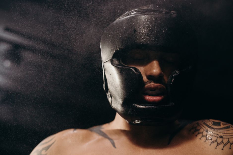 A muscular boxer wearing a head guard trains intensely in a dim gym setting.