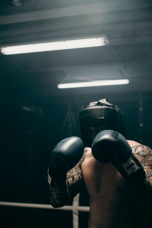 Free Shirtless boxer with tattoos preparing for a fight in a dimly lit gym, wearing protective gear. Stock Photo