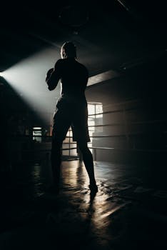 A silhouetted boxer practices in a gym with dramatic lighting and sun rays.