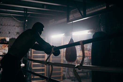 Moody silhouette of a boxer leaning in a dimly lit gym with punching bag and boxing ring.