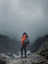 Woman in orange jacket standing on rocky mountain peak surrounded by fog.