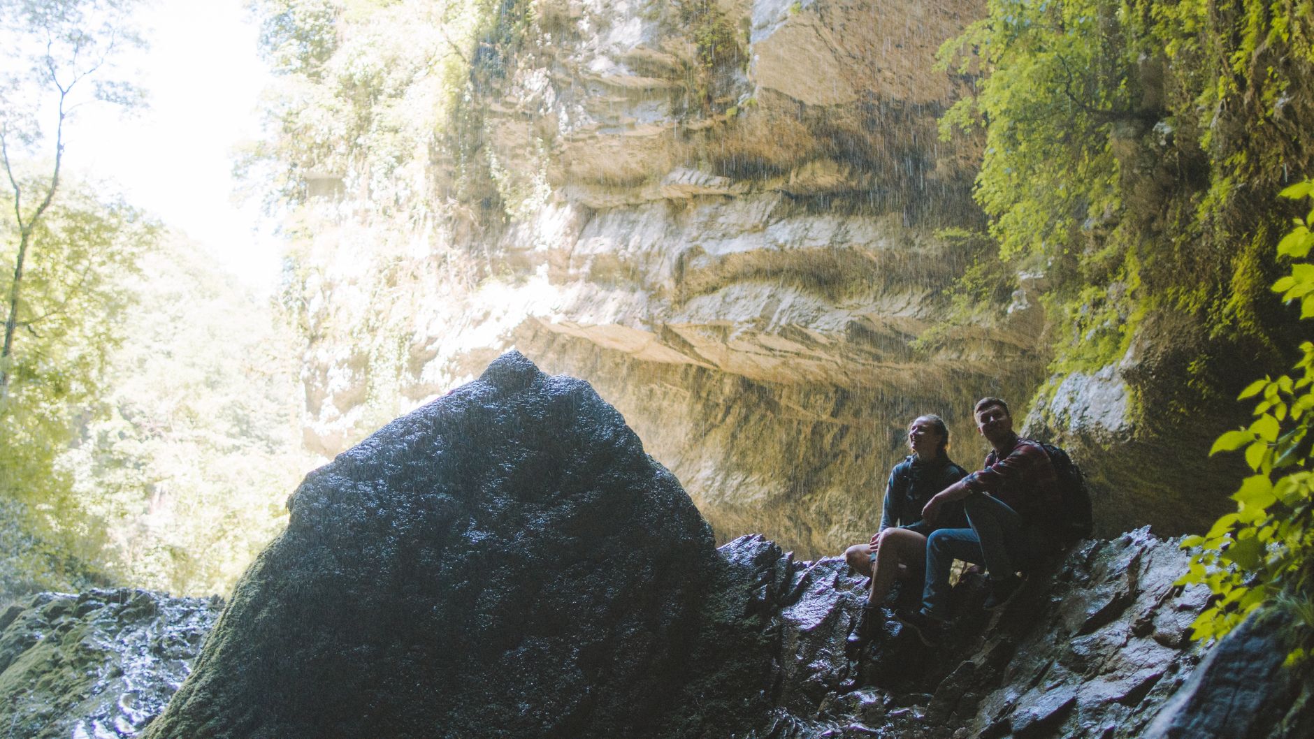 A couple sits in a scenic cave, enjoying the natural beauty surrounding them during a hiking adventure.