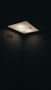 From below dirty ceiling lamp dimly shining corner of dark empty lift with ventilation holes in walls