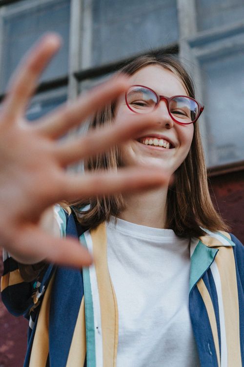Free Happy teenage girl with glasses smiling and waving outdoors in a playful moment. Stock Photo