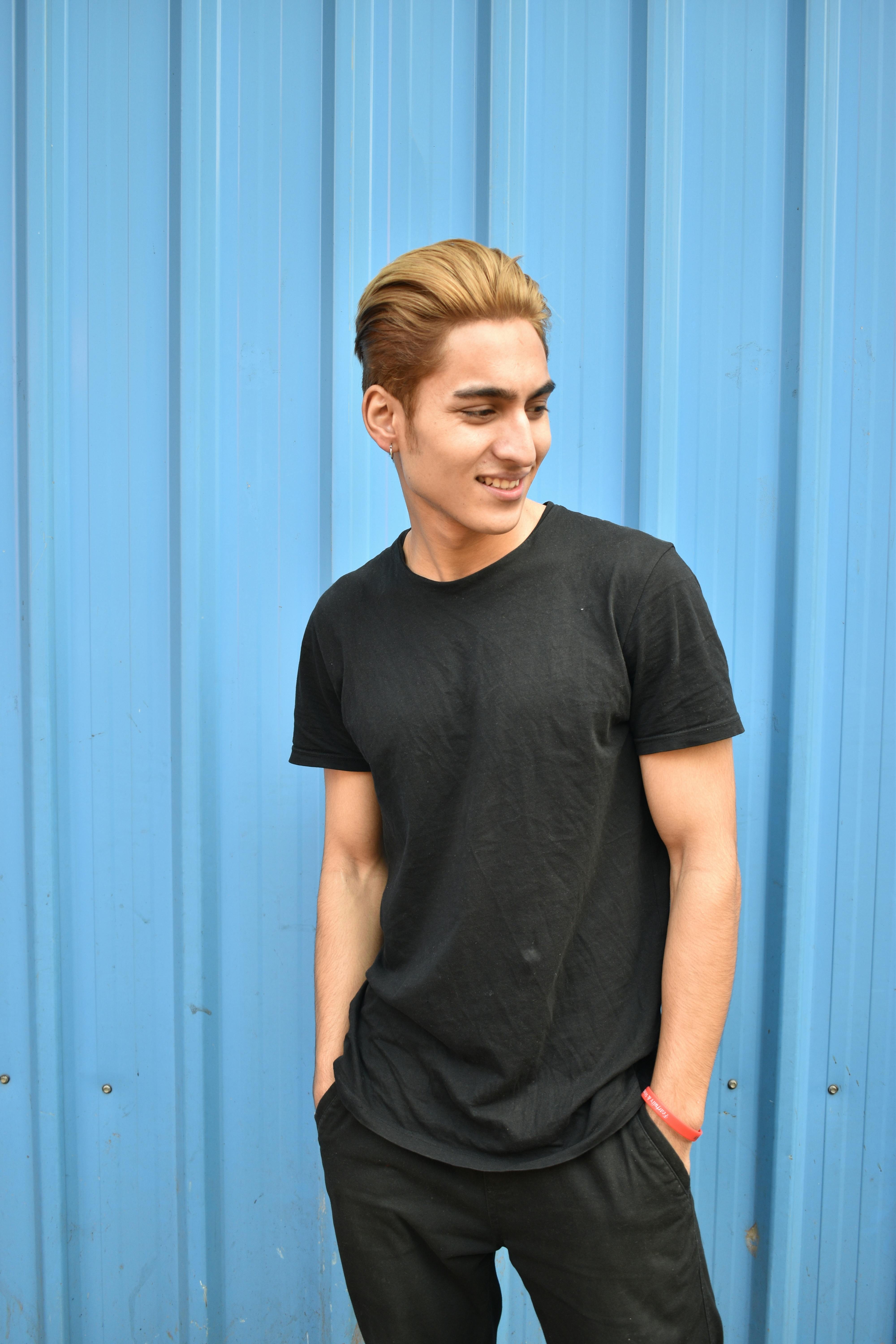 A young man smiles with hands in pockets, posing against a blue corrugated wall.