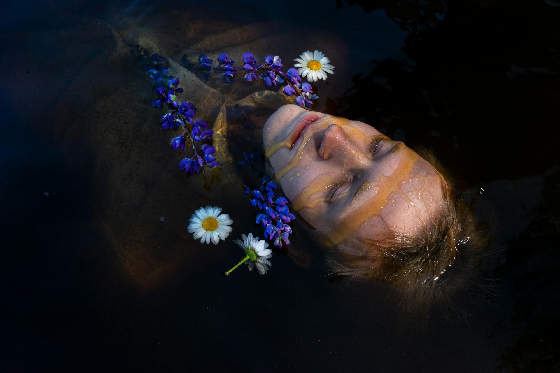 A serene portrait of a person floating with daisies and blue flowers in calm water.