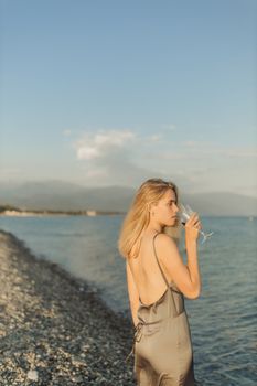 日落时分，一名身穿别致晚礼服的女子在海滩上品着葡萄酒
