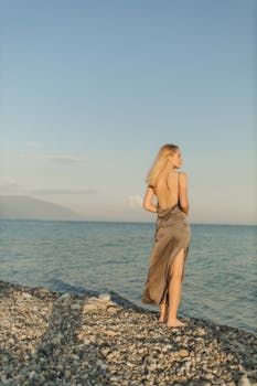 日落时分，一位优雅的女人站在岩石海滩上，凝视着宁静的大海