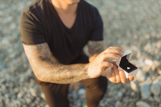 Man kneeling on a beach, presenting a diamond ring in a proposal scene.