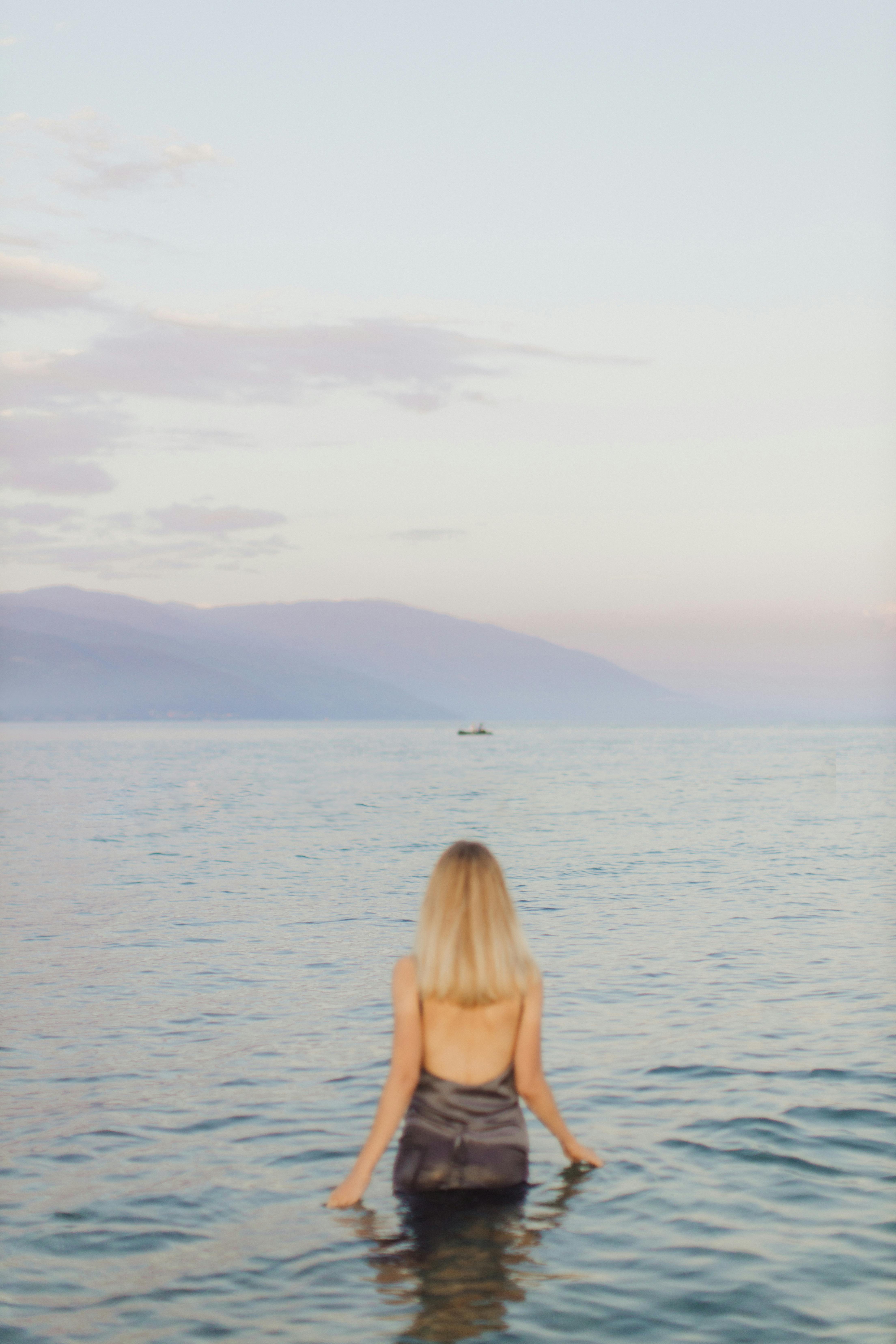 A serene woman stands waist-deep in calm waters, gazing towards distant mountains at dusk.