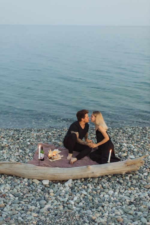 Free A couple sharing a romantic moment on a pebbly beach at sunset, surrounded by candles and wine. Stock Photo