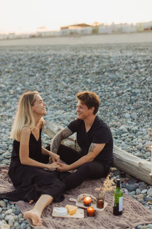 Free A couple enjoys a romantic picnic on a pebbled beach at sunset, sharing a moment together. Stock Photo