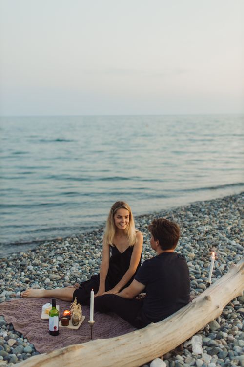 Free Couple enjoying a romantic picnic on a pebble beach at sunset with candles and wine. Stock Photo