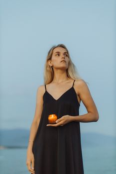日落时分，一位身着黑色连衣裙的安详女人手持点燃的蜡烛，站在海边