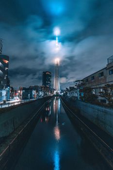 夜晚，东京天空树在雾气笼罩的城市景观中闪闪发光，倒映在一条反光的运河上