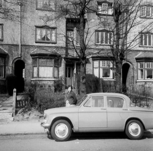 Free Grayscale Photo of Vintage Car Parked Along The Street With Woman Looking Stock Photo