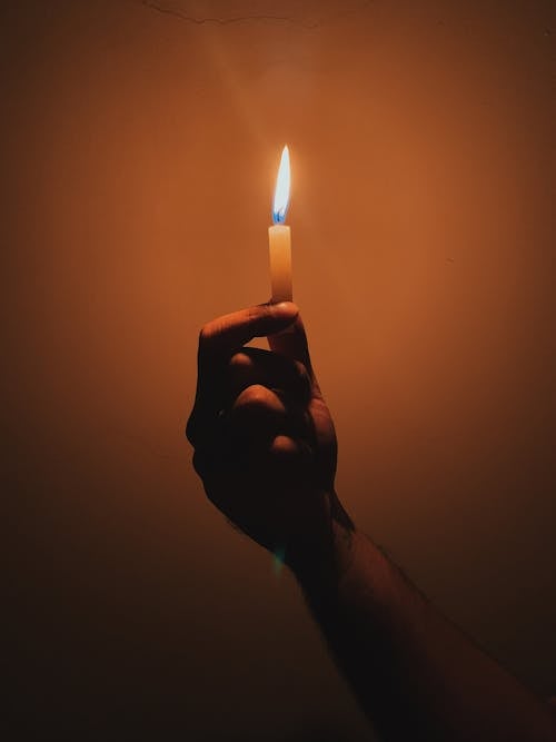 Crop anonymous person holding burning yellow candle in dark room with white background