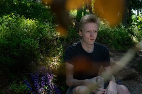 Pensive male in casual clothes smoking cigarette while sitting in forest in sunny summer day