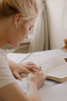 Focused young woman writing notes from an open textbook at a desk, creating a study environment.