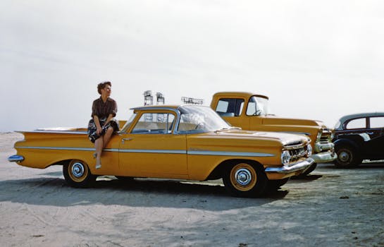 A woman sits on a vintage yellow car parked outdoors with classic vehicles around.