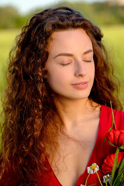 Free Peaceful young lady with ginger curly hair relaxing in blooming meadow with closed eyes on sunny day Stock Photo