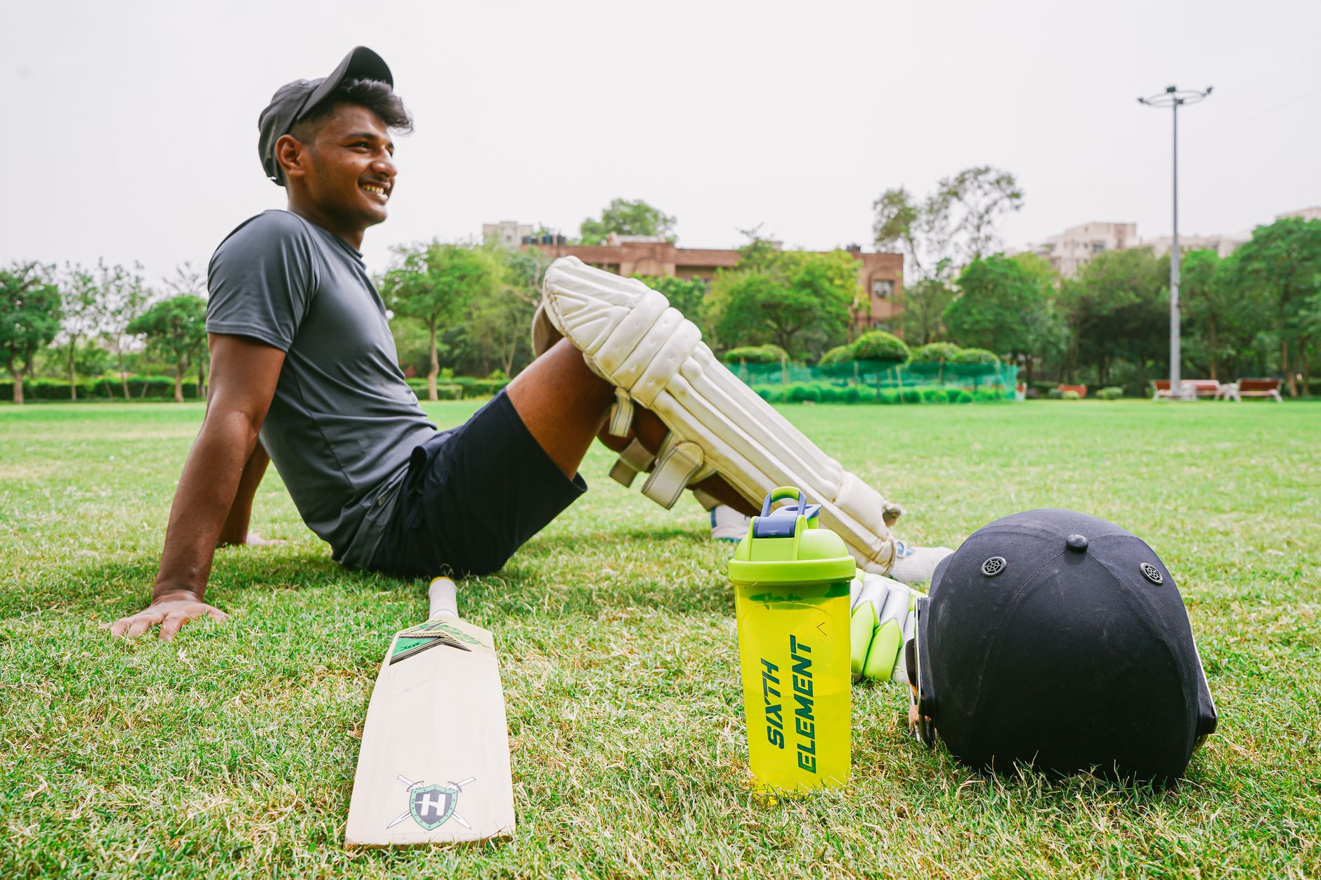 Side view of full body happy sportsman wearing sportswear and cricket pads resting on lawn beside cricket bat and helmet and sport water bottle
