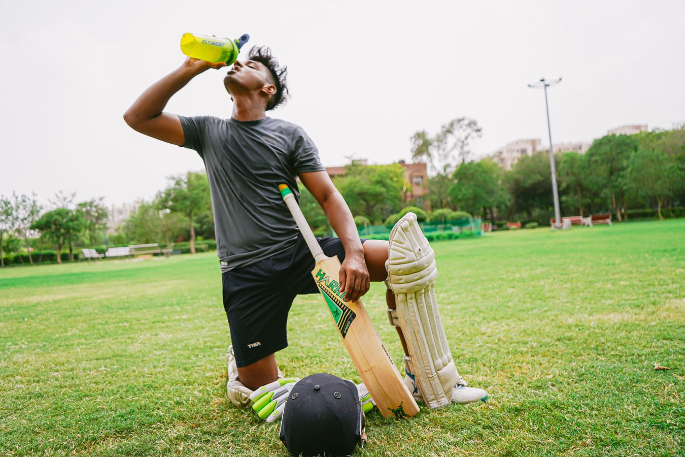 Indian cricket player kneeling on grass and drinking water from sports bottle mid-game