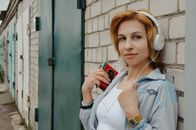 Charming red haired female listening to music in headphones while standing near brick building and looking away
