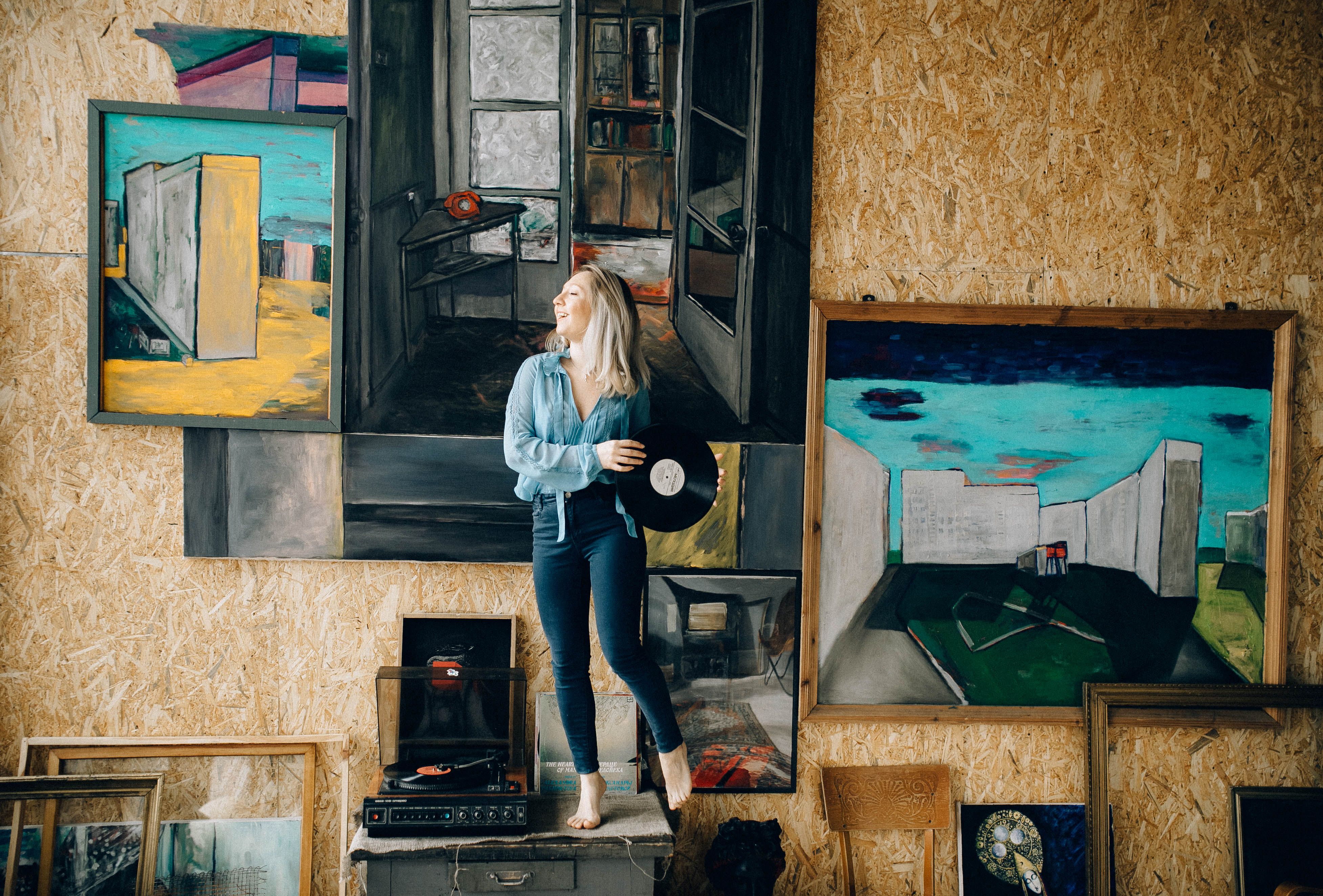 A woman holding a vinyl record stands amidst eclectic artwork and retro decor.