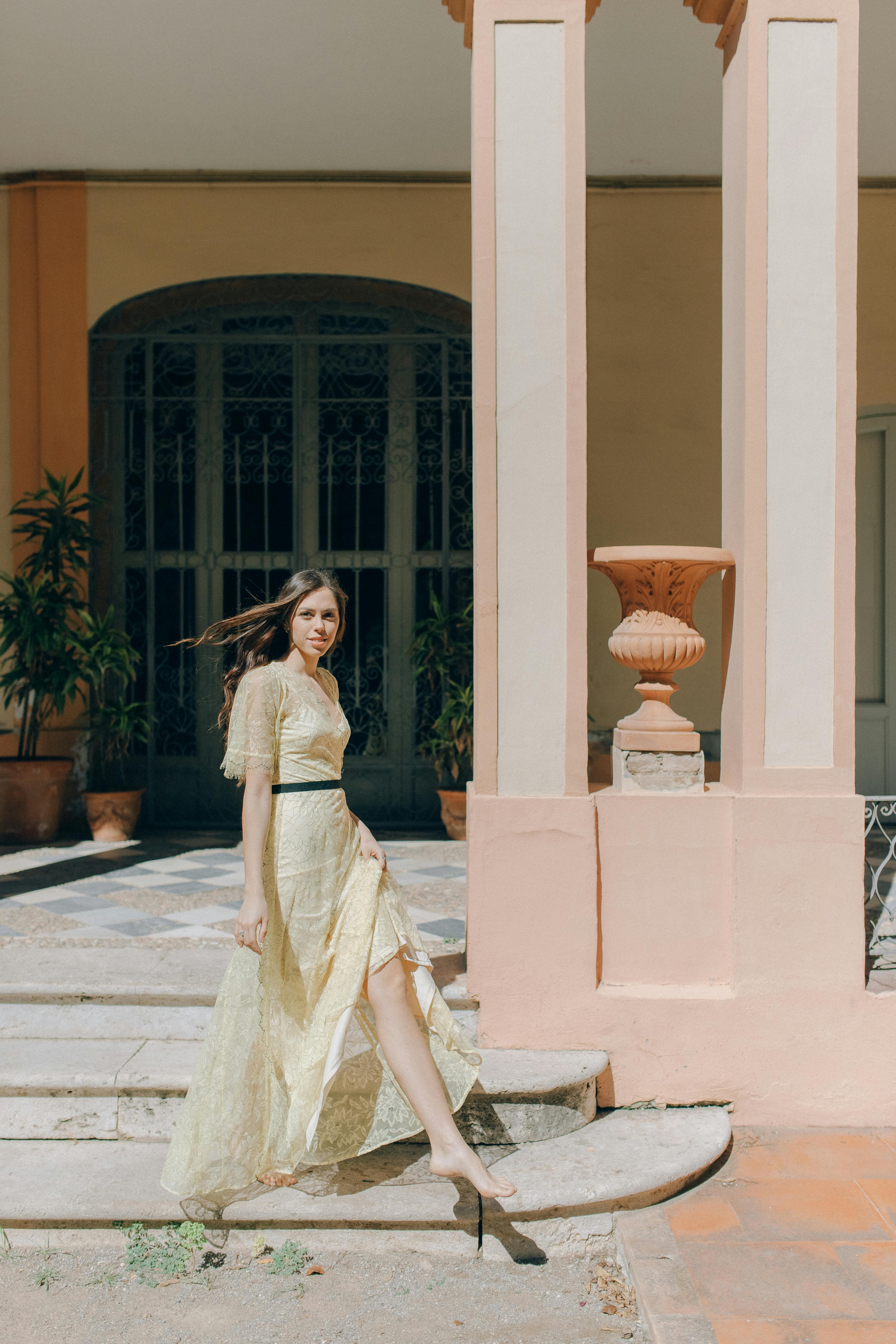 A woman in a long lace dress elegantly poses on steps outdoors under the sun.