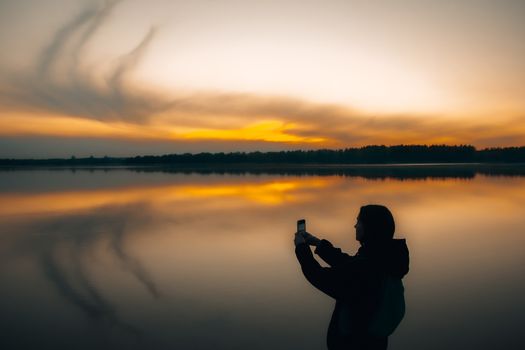 A silhouette of a person taking a photo by a calm lake during sunset, reflecting vibrant skies.