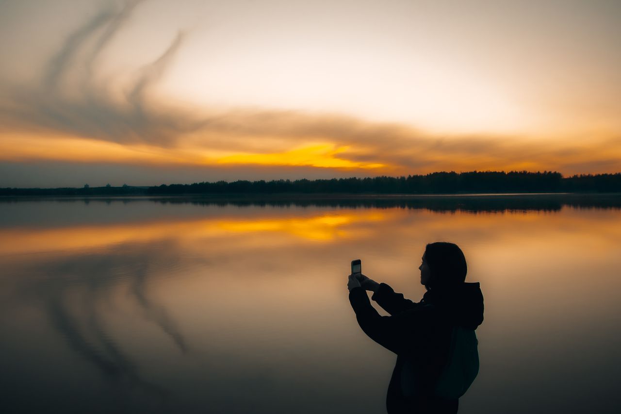 a silhouette of a person taking a photo by a calm lake during sunset r