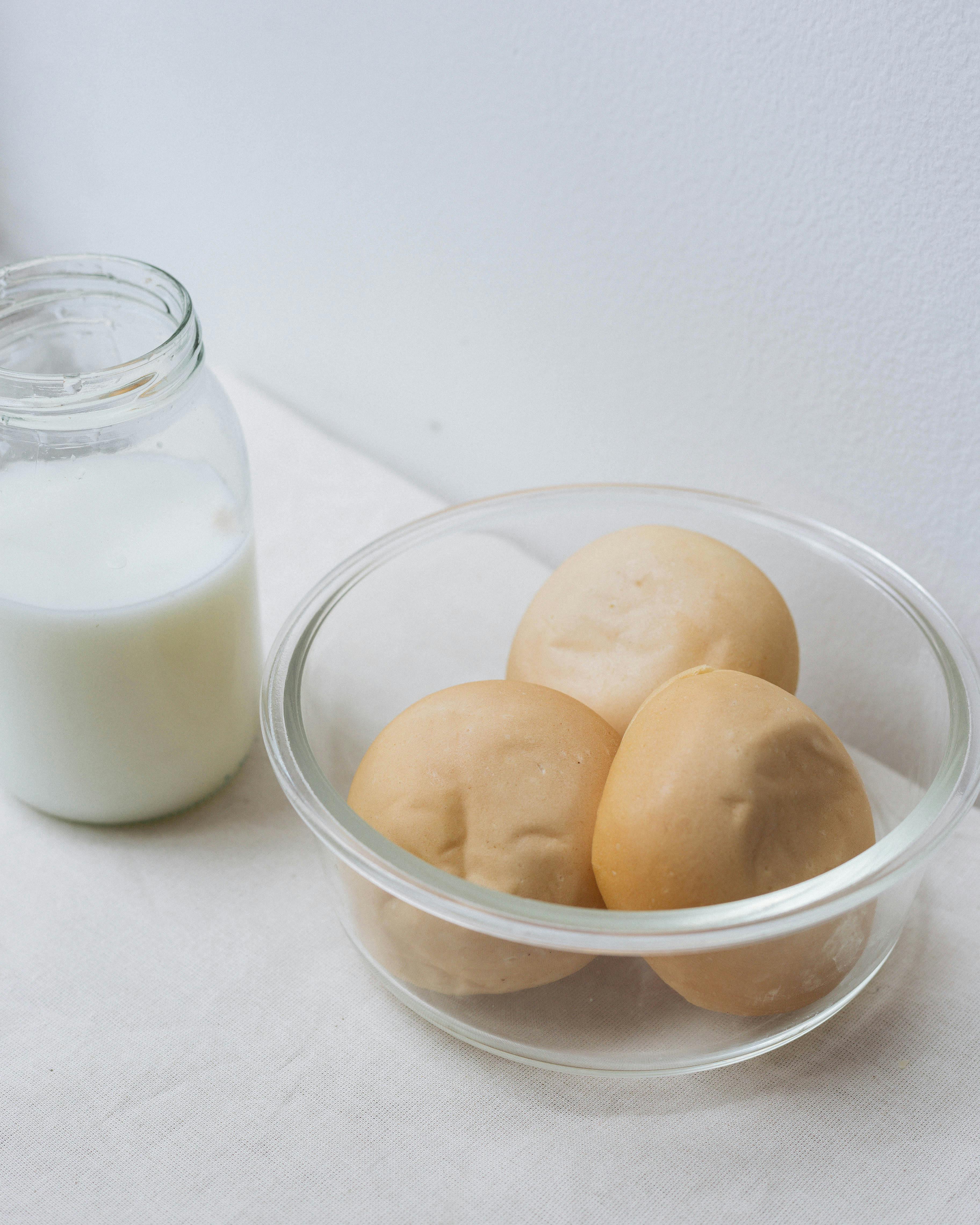 A glass bowl of freshly baked rolls next to a jar of milk on a light table setting.