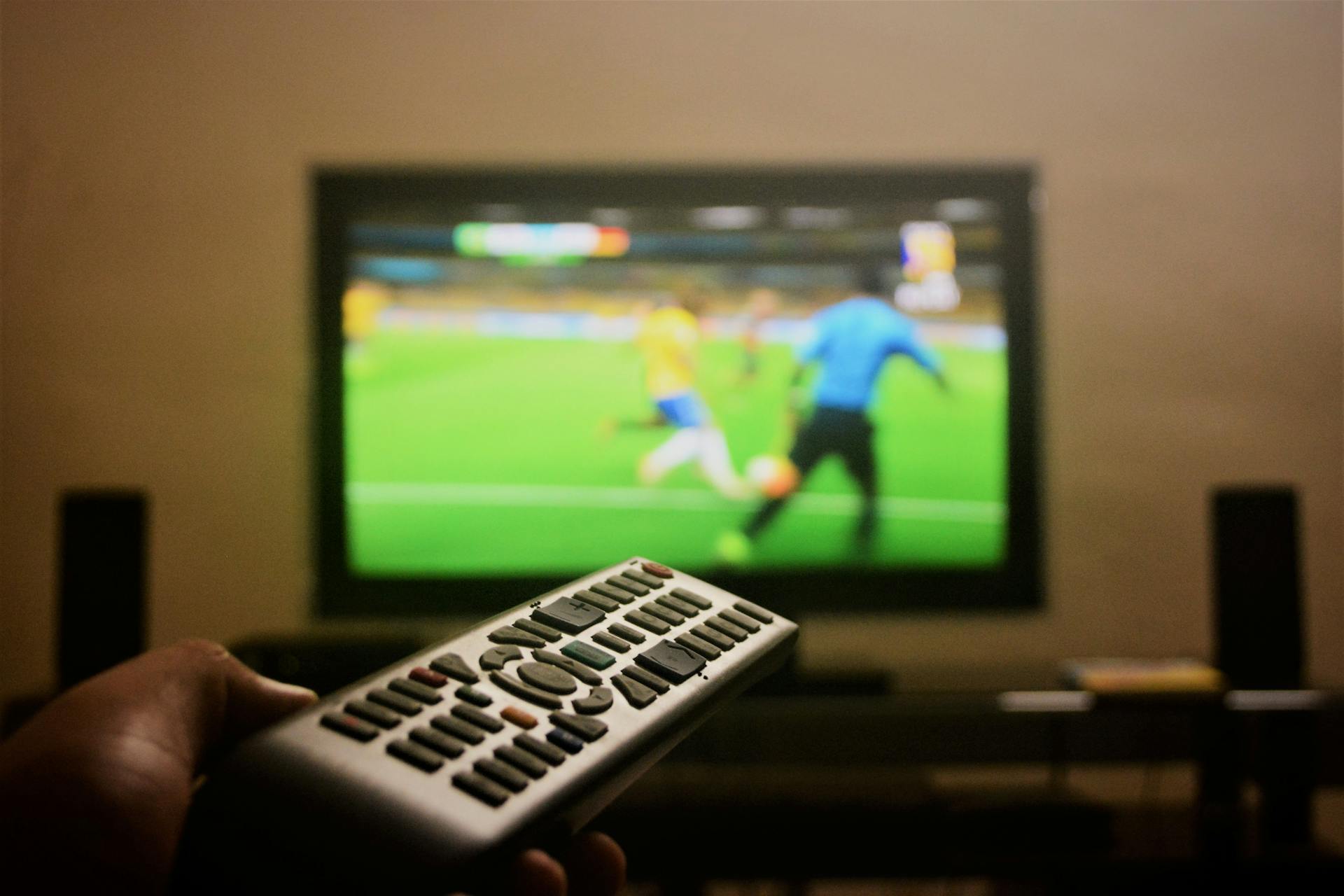 Close-up of a hand holding a remote control in front of a TV showing a football match.