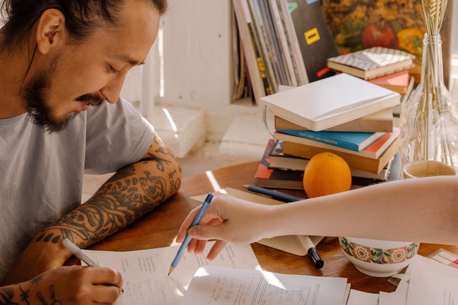 A tattooed Asian student concentrates on studying with books and papers at a desk.