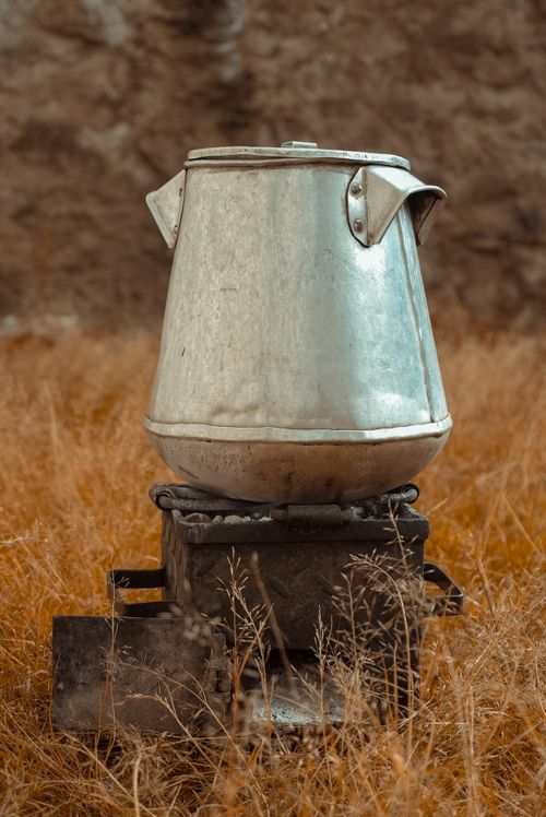 Free Close-up of an antique metal kettle on a rustic stove amidst dry grass outdoors. Stock Photo