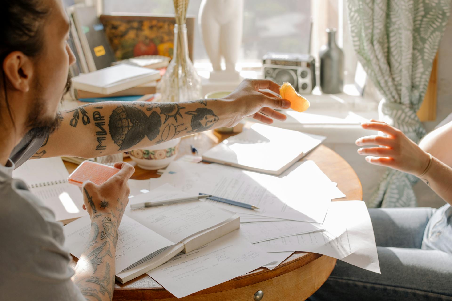 Tattooed students exchanging notes during a study session with papers spread on a round table.