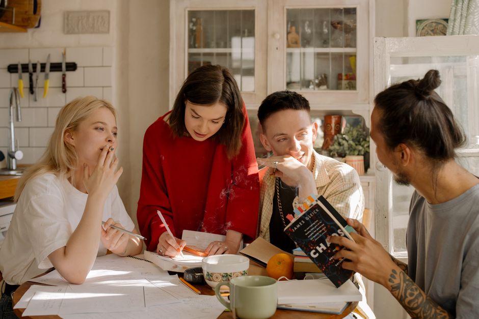 Group of young adults studying together at a kitchen table filled with books and notes.