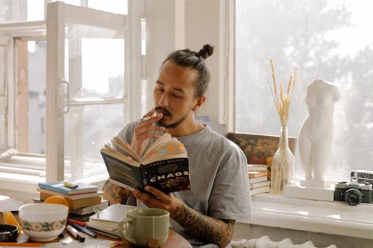 Young man reading and smoking by a window with books and decor, enjoying a relaxed atmosphere.